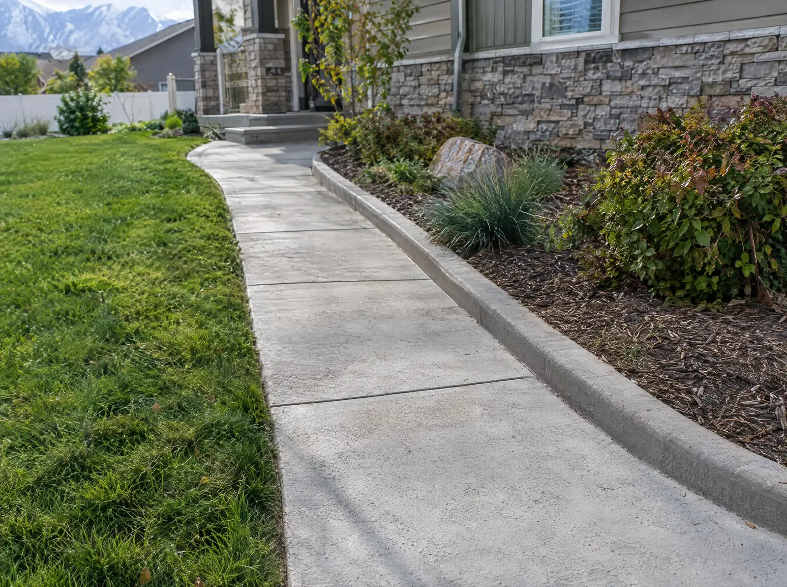 Concrete walkway curving up a slope beside a cut lawn, a mulched bed of shrubs and a stone-clad house.