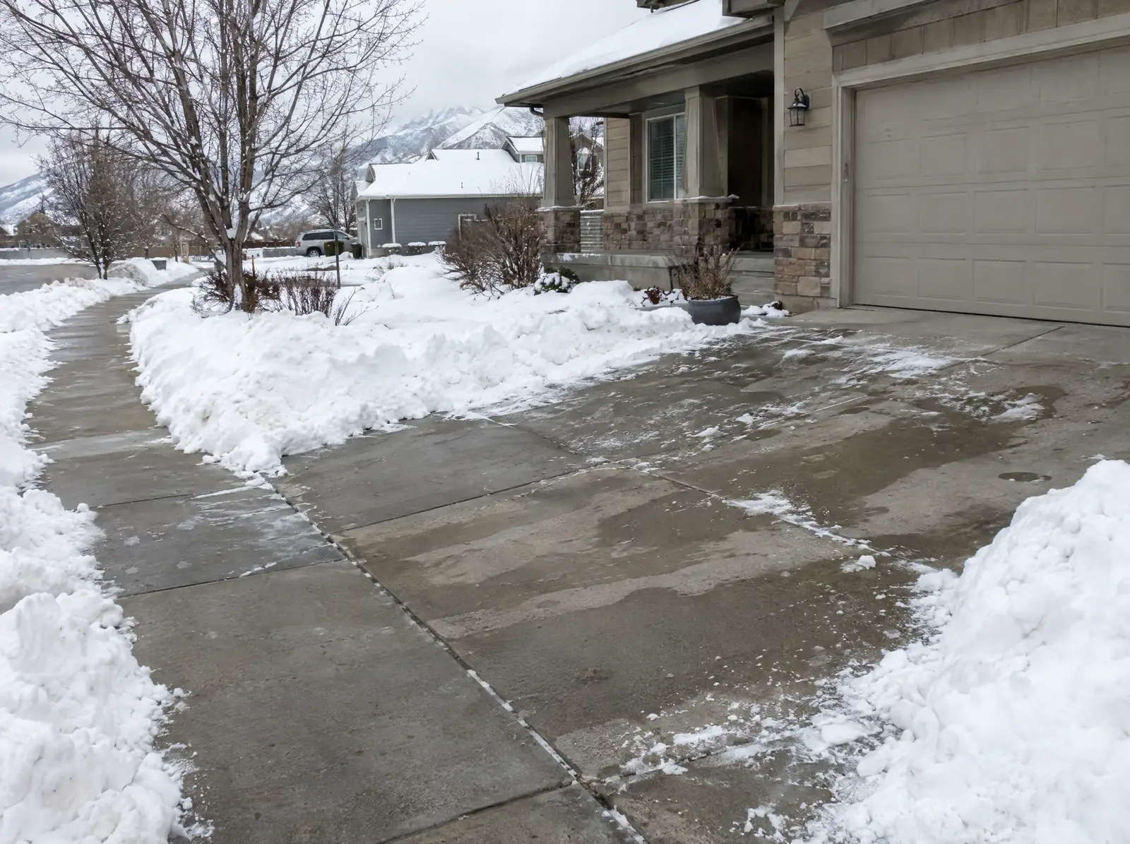 Concrete driveway and walk cleared down to the slab between banks of snow, with a bare tree and the house behind.